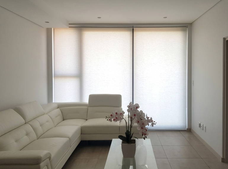 A wide interior view of automated roller blinds with light filtering fabric installed in a modern Johannesburg home with a clean cream leather sectional couch.